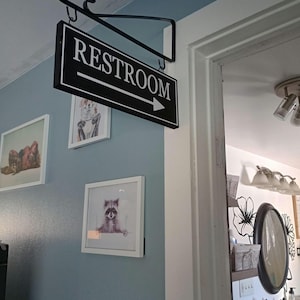 May include: Black and white sign with the word "RESTROOM" and an arrow pointing to the right. The sign is hanging from a black metal hook.