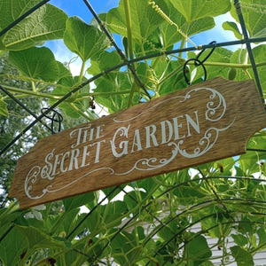 Puede incluir: Un letrero de madera rectangular con bordes decorativos colgado de un enrejado de alambre metálico. El letrero presenta el texto blanco THE SECRET GARDEN en una fuente estilizada con adornos, suspendido por dos anillos de metal negro.