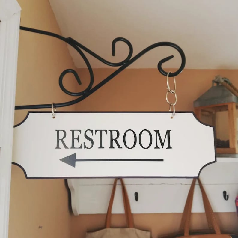 May include: White sign with black lettering that reads "RESTROOM" with a black arrow pointing to the left. The sign is hanging from a black metal hook.