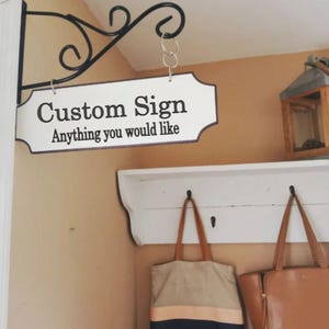 Puede incluir: Un letrero blanco con letras negras que dice "Custom Sign Anything you would like" colgado de un gancho de metal negro. El letrero está colgado sobre un estante blanco con dos ganchos y dos bolsas colgadas de los ganchos.
