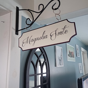 May include: A black metal sign holder with a white sign that reads "Magnolia Suite" in cursive script. The sign is hanging on a wall with a light blue paint color.