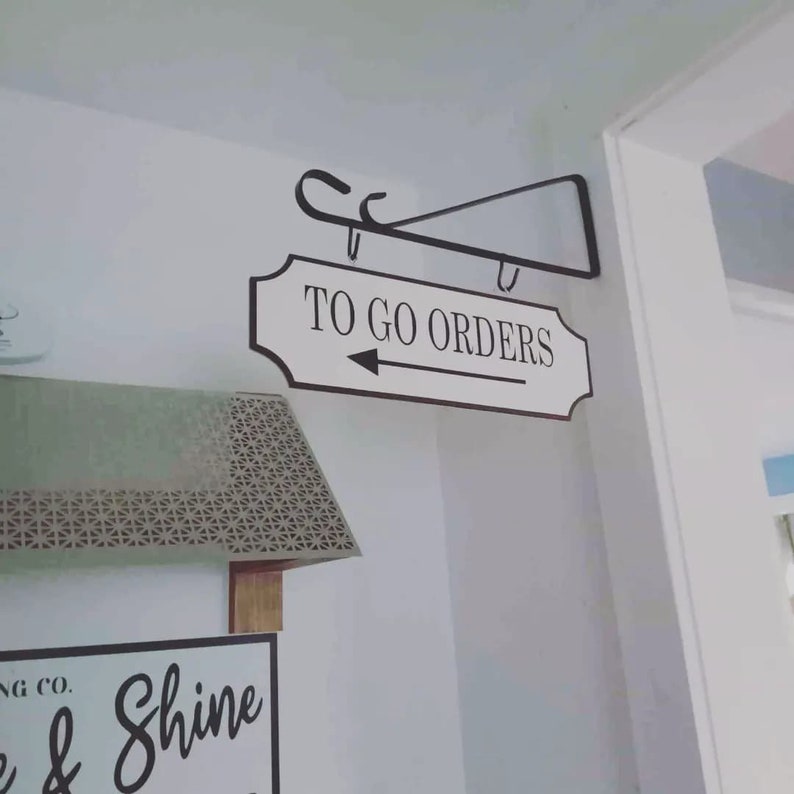 May include: A white sign with black text that reads "TO GO ORDERS" with a black arrow pointing to the right. The sign is hanging from a black metal bracket.