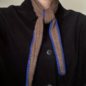 May include: A brown knitted scarf with blue trim. The scarf is tied around the neck in a loose knot.