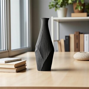 May include: A black, textured vase with a geometric design. The vase has a narrow neck and a wide base, and it is sitting on a light-colored wooden table. The vase is approximately 12 inches tall.