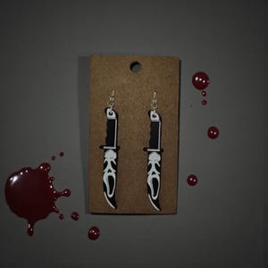 May include: A pair of novelty earrings featuring a black and white knife design with a Scream mask. The earrings are displayed on a brown card with a hole at the top. Red paint splatters are scattered around the earrings.