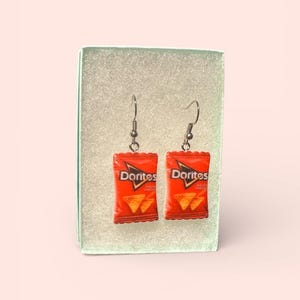 May include: A pair of novelty earrings featuring miniature red Doritos nacho cheese chip bags. The earrings have silver-colored fish hook ear wires and are displayed inside a small, light green box.