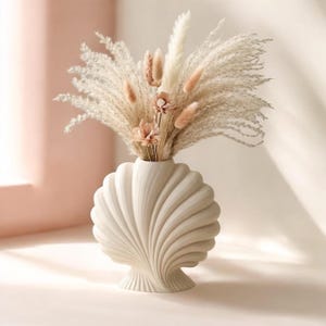 May include: A cream-colored, seashell-shaped vase holds a bouquet of dried pampas grass and delicate, light pink flowers. The vase has a textured, ridged design and sits on a neutral surface, creating a soft, natural aesthetic.