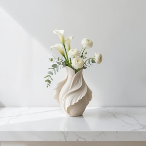 May include: A cream-colored, sculptural vase filled with white calla lilies, ranunculus, and eucalyptus. The vase has a unique, flowing design, resembling petals. The arrangement sits on a white surface, creating a clean and elegant aesthetic.