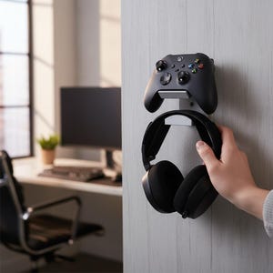Puede incluir: Un mando Xbox negro y unos auriculares negros están montados en un soporte de pared. El mando está colocado encima de los auriculares. El soporte es plateado y está fijado a una pared gris clara. El fondo muestra un escritorio con un ordenador y una ventana.
