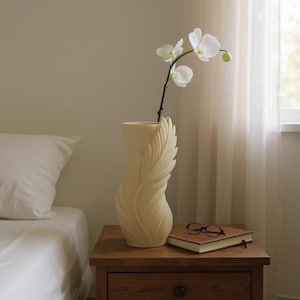 May include: A cream-colored vase with a feather design holds a white orchid. The vase sits on a wooden nightstand next to a closed book and eyeglasses. A white pillow and bed are in the background.