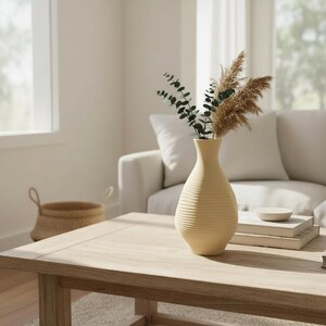 May include: A cream-colored, ribbed ceramic vase holding dried pampas grass and eucalyptus sprigs sits on a light wood coffee table. The vase is the focal point, with a neutral-toned living room in the background.