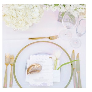 May include: A white table setting with a gold rimmed plate, gold silverware, and a white napkin. A white tulip is in a small white vase on the napkin. A gold seashell is on the napkin with the name "Ann Rose" written on it.