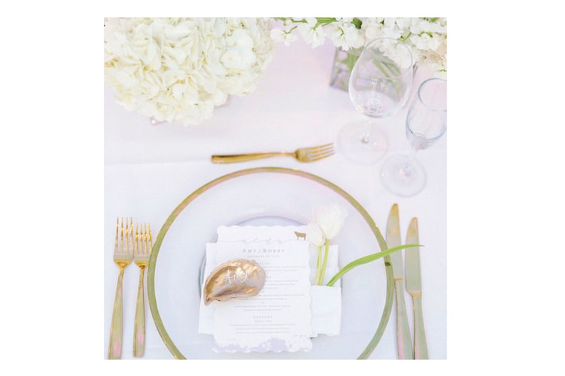 May include: A white table setting with a gold rimmed plate, gold silverware, and a white napkin. A white tulip is in a small white vase on the napkin. A gold seashell is on the napkin with the name "Ann Rose" written on it.