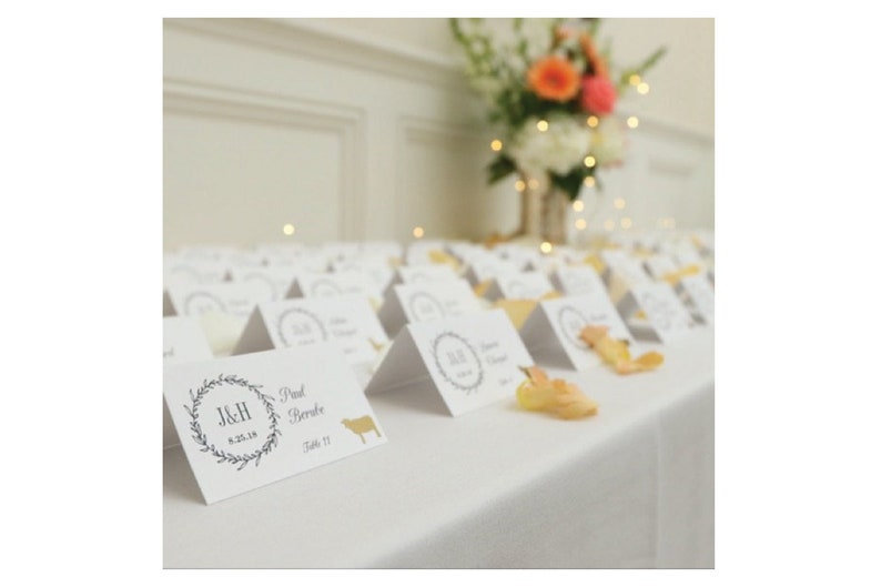 May include: White table with place cards for a wedding reception. The cards are folded into a triangle shape and have a floral wreath design with the initials J&H and the date 8.25.18. The text "Paul Berube" and "Table 11" are also printed on the card.
