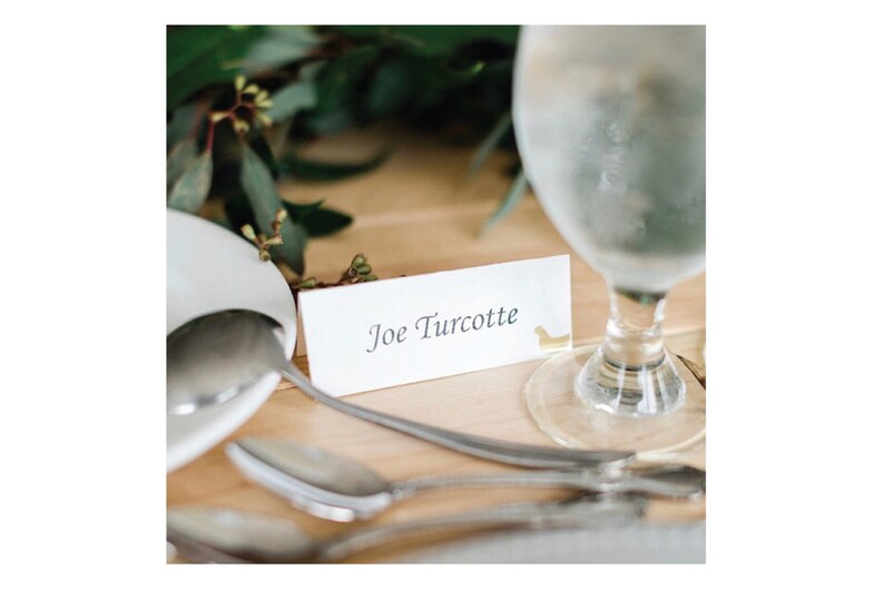 May include: A white place card with the name "Joe Turcotte" written in black ink. A small gold figurine is attached to the right side of the card.