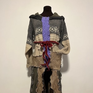 May include: A patchwork hooded coat in shades of gray, beige, and brown, with a purple zipper and a red belt. The coat features various knit and woven textures, including cable knit and snowflake patterns. The coat is displayed on a mannequin.