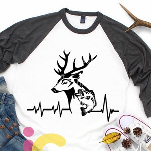 May include: A white and gray baseball tee shirt with a black silhouette of a deer head with a fish in its mouth and a heartbeat graphic below. The shirt is on a white surface with blue jeans, fall leaves, and white sneakers.
