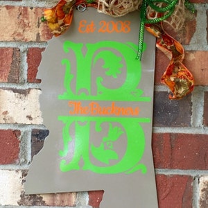May include: A gray metal sign in the shape of Mississippi, with the text "Est 2008" in orange. The sign features the name "The Buckners" and large green initials. The sign is decorated with fall-themed ribbons and twine.