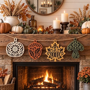 Puede incluir: Una guirnalda decorativa con cuatro adornos de madera colgantes con formas otoñales: una calabaza clara, una bellota naranja, una hoja de arce amarilla con una calabaza recortada y un hongo verde oscuro, suspendidos en un cordel marrón.