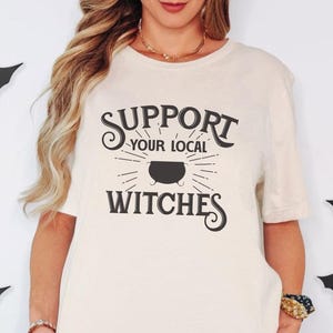 May include: A cream-colored short-sleeve t-shirt featuring the black text "SUPPORT YOUR LOCAL WITCHES" and a graphic of a cauldron. The shirt is paired with black shorts. Several black paper bat cutouts are positioned around the garment.
