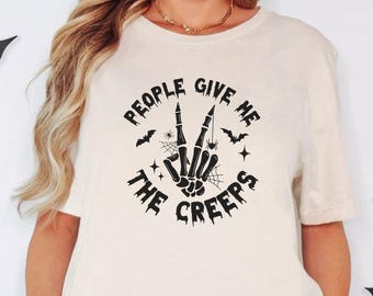 Puede incluir: Una camiseta de color crema con un gráfico negro de una mano de esqueleto haciendo el signo de la paz. El texto "PEOPLE GIVE ME THE CREEPS" está impreso en una fuente desgastada alrededor de la mano, junto a iconos de telarañas y murciélagos.
