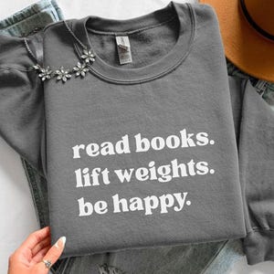 May include: A dark gray crewneck sweatshirt featuring the white printed text read books. lift weights. be happy. The garment is displayed alongside a pair of light blue denim jeans, a brown felt hat, and a silver-toned floral necklace.