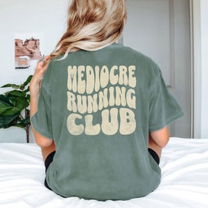 Puede incluir: Una camiseta verde salvia con las palabras "Mediocre Running Club" en una fuente retro de color crema. La camiseta es de corte holgado y parece estar hecha de un material suave. Se ve la parte trasera de la camiseta.