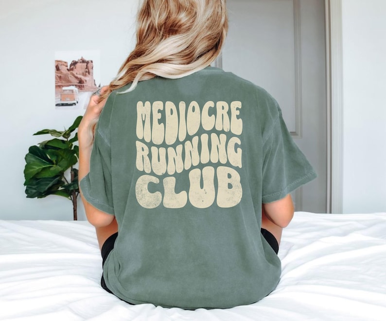 Puede incluir: Una camiseta verde salvia con las palabras "Mediocre Running Club" en una fuente retro de color crema. La camiseta es de corte holgado y parece estar hecha de un material suave. Se ve la parte trasera de la camiseta.