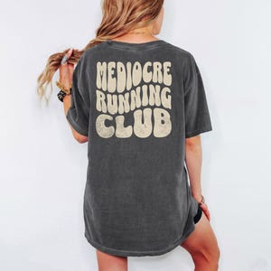 Puede incluir: Camiseta gris oscuro con el texto "Mediocre Running Club" en una fuente retro en la espalda. La camiseta tiene un aspecto ligeramente descolorido, lo que sugiere un aspecto vintage o desgastado. La camiseta es de estilo casual.