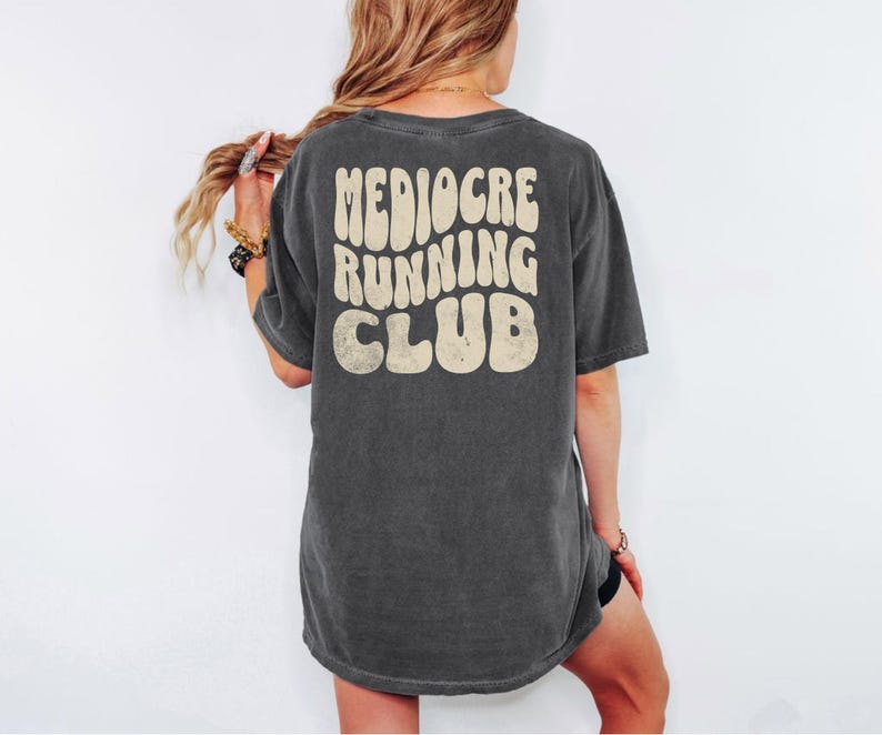 Puede incluir: Camiseta gris oscuro con el texto "Mediocre Running Club" en una fuente retro en la espalda. La camiseta tiene un aspecto ligeramente descolorido, lo que sugiere un aspecto vintage o desgastado. La camiseta es de estilo casual.
