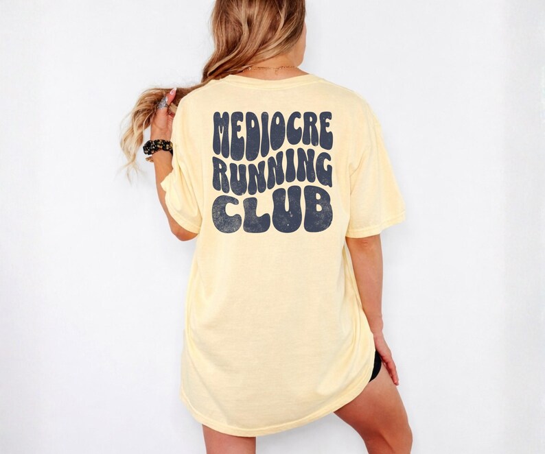 Puede incluir: Camiseta amarillo claro con la frase "Mediocre Running Club" impresa en azul oscuro, con letras de estilo retro. La camiseta tiene un corte holgado y casual.