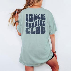 Puede incluir: Una camiseta verde claro con las palabras "Mediocre Running Club" en letras retro de color azul oscuro. La camiseta es de gran tama&ntilde;o y tiene mangas cortas. La persona lleva un pantal&oacute;n corto negro.