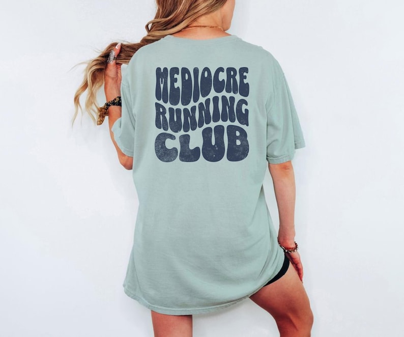 Puede incluir: Una camiseta verde claro con las palabras "Mediocre Running Club" en letras retro de color azul oscuro. La camiseta es de gran tama&ntilde;o y tiene mangas cortas. La persona lleva un pantal&oacute;n corto negro.