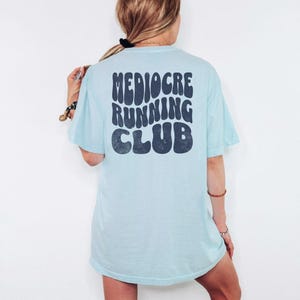 Puede incluir: Camiseta azul claro con el texto "Mediocre Running Club" en azul oscuro, con una fuente retro. La camiseta es de corte holgado y tiene mangas cortas. La imagen muestra la parte trasera de la camiseta.