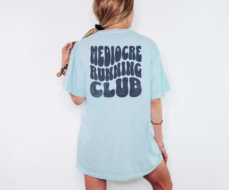 Puede incluir: Camiseta azul claro con el texto "Mediocre Running Club" en azul oscuro, con una fuente retro. La camiseta es de corte holgado y tiene mangas cortas. La imagen muestra la parte trasera de la camiseta.