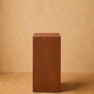 May include: A tall, rectangular pedestal with a weathered, rusted metal finish. The object features a flat top surface and four vertical sides, displaying a consistent, textured, reddish-brown oxidised appearance throughout its entire structure.
