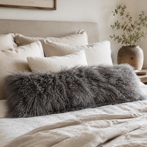 Peut inclure: Un long coussin lombaire rectangulaire recouvert d'une fourrure gris foncé à la texture bouclée et hirsute. Le coussin est placé horizontalement devant un lit superposé de plusieurs oreillers carrés et rectangulaires beige clair.