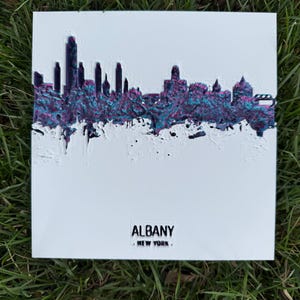 May include: A white square art piece featuring the Albany, New York skyline. The skyline is rendered in a dark purple and teal color scheme. The word "ALBANY" and "NEW YORK" are printed in black at the bottom.