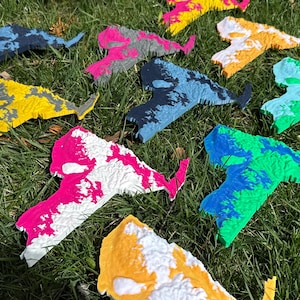 May include: A collection of colorful, textured, abstract shapes resembling a map outline, scattered on green grass. The shapes are painted in various colors, including yellow, pink, blue, and green, with white accents. The overall theme is artistic and decorative.