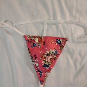 May include: A pink floral thong with white straps. The thong features a triangular front panel with a pattern of red, white, and blue flowers. The straps are thin and white, designed to be discreet.