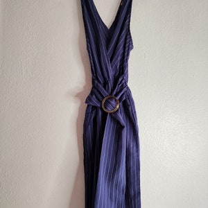 May include: A sleeveless navy blue jumpsuit with white pinstripes, hanging on a clear plastic hanger. The jumpsuit features a V-neckline, a fabric belt, and a large, round, wooden buckle. The fabric is gathered at the waist.