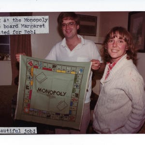 May include: A handmade Monopoly game board, knitted in green with red, yellow, and black details. The board is held by two people. Text on the board reads "MONOPOLY". Text above reads "Look at the Monopoly game board Margaret knitted for Rob!" and below reads "Beautiful job!"