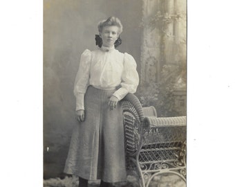 Pretty young woman with a large bow in her hair leaning on an elaborate wicker chair. Undated vintage RPPC.