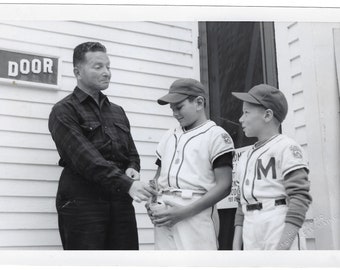May include: Black and white photograph of a man and two young baseball players. The man is handing a bottle to one of the boys. The boys are wearing baseball uniforms and caps. The background is a white building with a door.