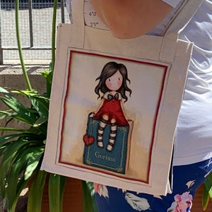 May include: A white canvas tote bag with a colorful illustration of a girl sitting on a book. The book is blue with a red heart and the word "Gorjuss" printed on it.