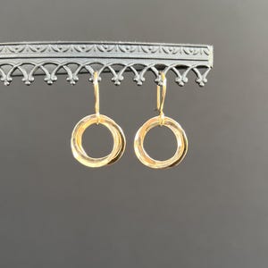 May include: A pair of gold-coloured hoop earrings. Each earring has a simple, circular design. The earrings are displayed on a decorative black metal stand against a grey background. The earrings are suspended from a hook.