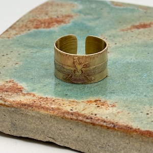 May include: A gold-colored adjustable ring with an engraved design. The ring features a detailed pattern around the band, and is displayed on a textured, multi-colored surface. The ring's design is centered on the band.