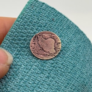 Può includere: Una piccola moneta rotonda color rame con una mappa dettagliata di un paese. La moneta è appoggiata su un tessuto turchese strutturato, creando un contrasto di colore e consistenza. La superficie della moneta ha un aspetto leggermente invecchiato.