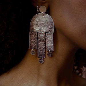May include: Silver dangle earrings with a large, semi-circular charm featuring intricate calligraphy. The charm is attached to five smaller rectangular charms, each with calligraphy, and four small round charms.