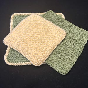 May include: Three handmade crochet washcloths. One is a light yellow, and two are green. The washcloths are square with a textured stitch pattern and a contrasting border. The washcloths are likely for the kitchen or bathroom.